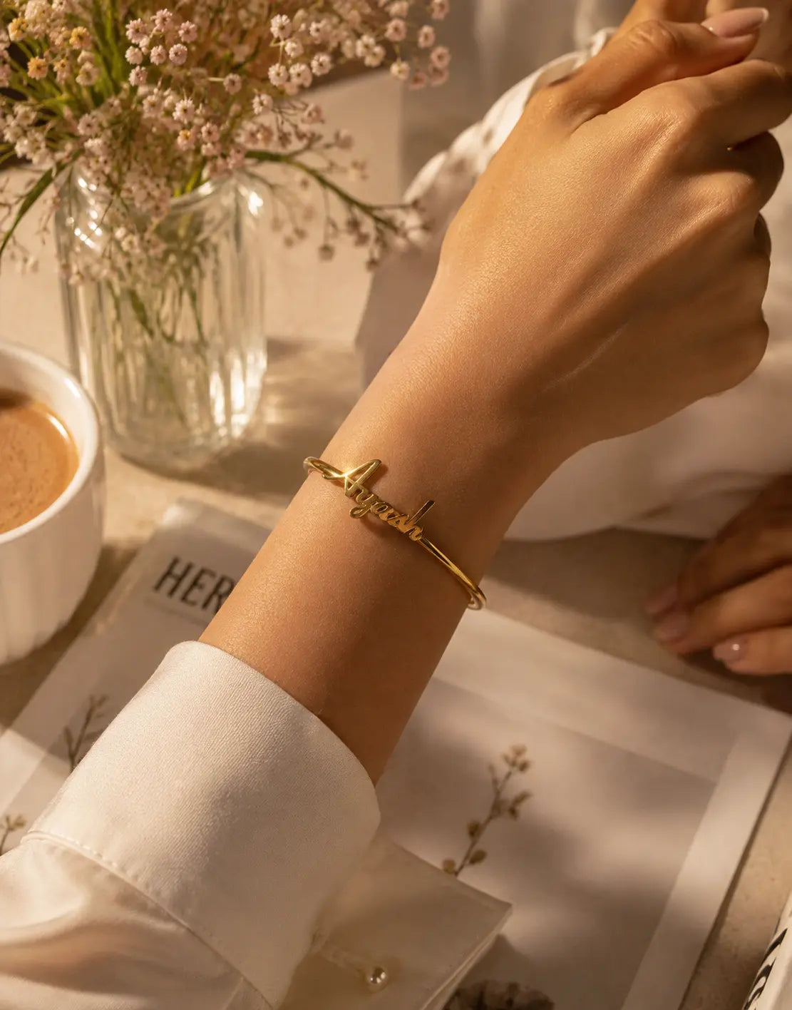 A gold-plated metal cuff bracelet with a small script charm, worn on a person's wrist. The scene is styled with a white shirt cuff, a glass vase of baby's breath flowers, and a cup of coffee on a table.
