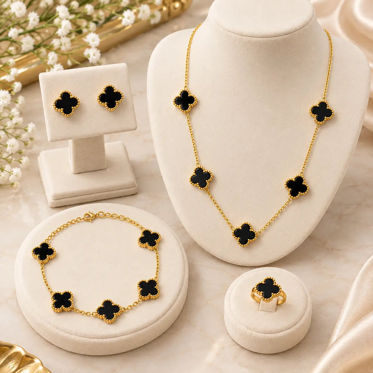 Gold jewelry set with black clover motifs, displayed on cream jewelry stands and a ring box. The set includes a necklace, bracelet, earrings, and ring, arranged on a light marble surface with soft decorative flowers in the background.