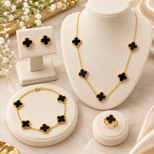 Gold jewelry set with black clover motifs, displayed on cream jewelry stands and a ring box. The set includes a necklace, bracelet, earrings, and ring, arranged on a light marble surface with soft decorative flowers in the background.
