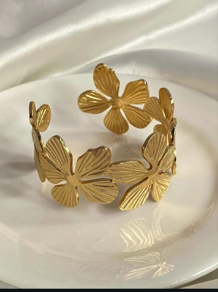 A gold-tone open cuff bracelet with textured, ribbed butterfly or flower-like links, displayed on a white ceramic plate against a draped white satin background.