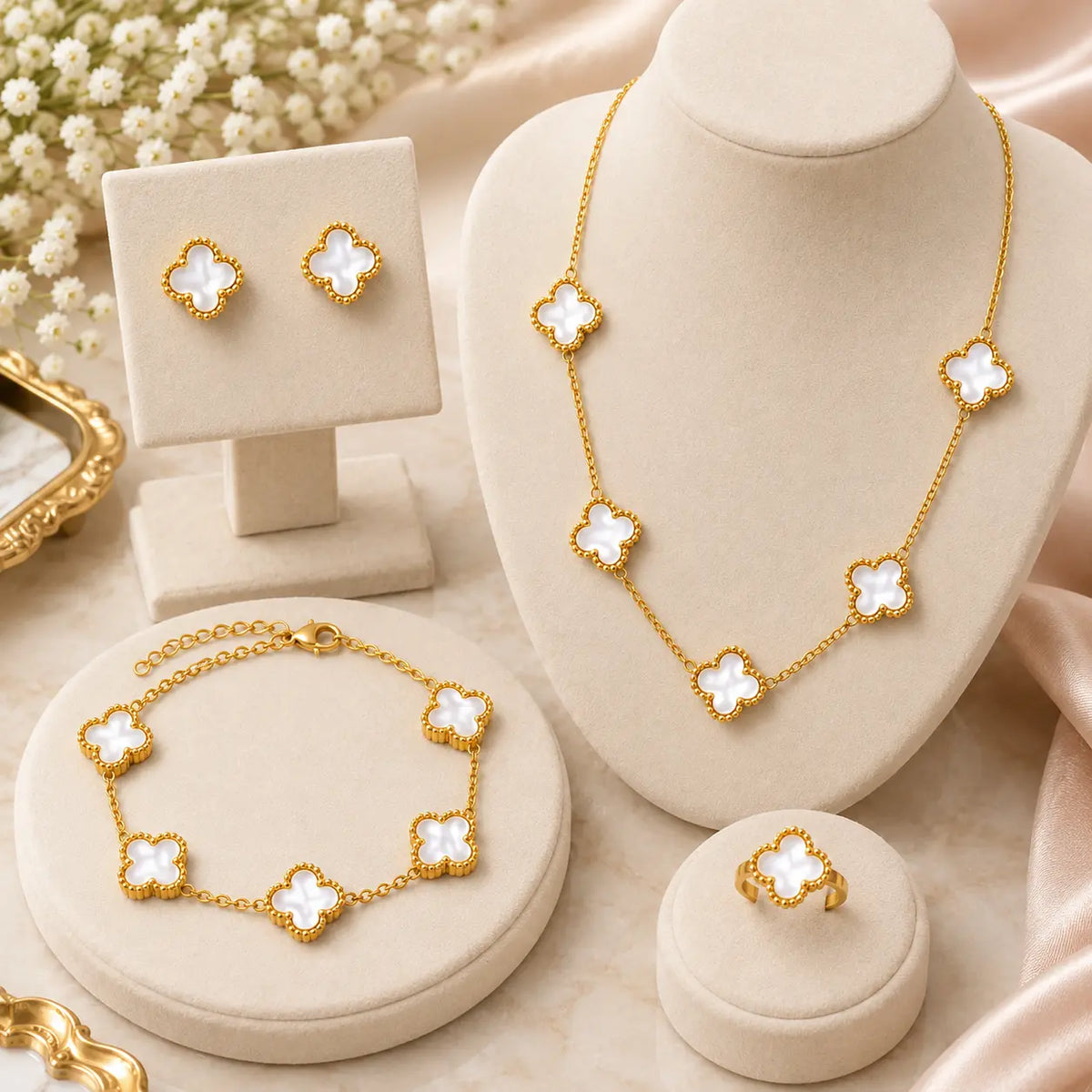 Gold jewelry set with white clover motifs, including a necklace, bracelet, ring, and matching earrings, displayed on cream-colored jewelry stands and a satin fabric background.